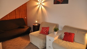 A cozy living room with a brown sofa, a beige sofa, and a small table with a lamp. There is a plant on the table and a red pillow on the beige sofa.