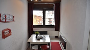 A small, cozy room with a table and bench, a window with curtains, and a view of buildings outside.