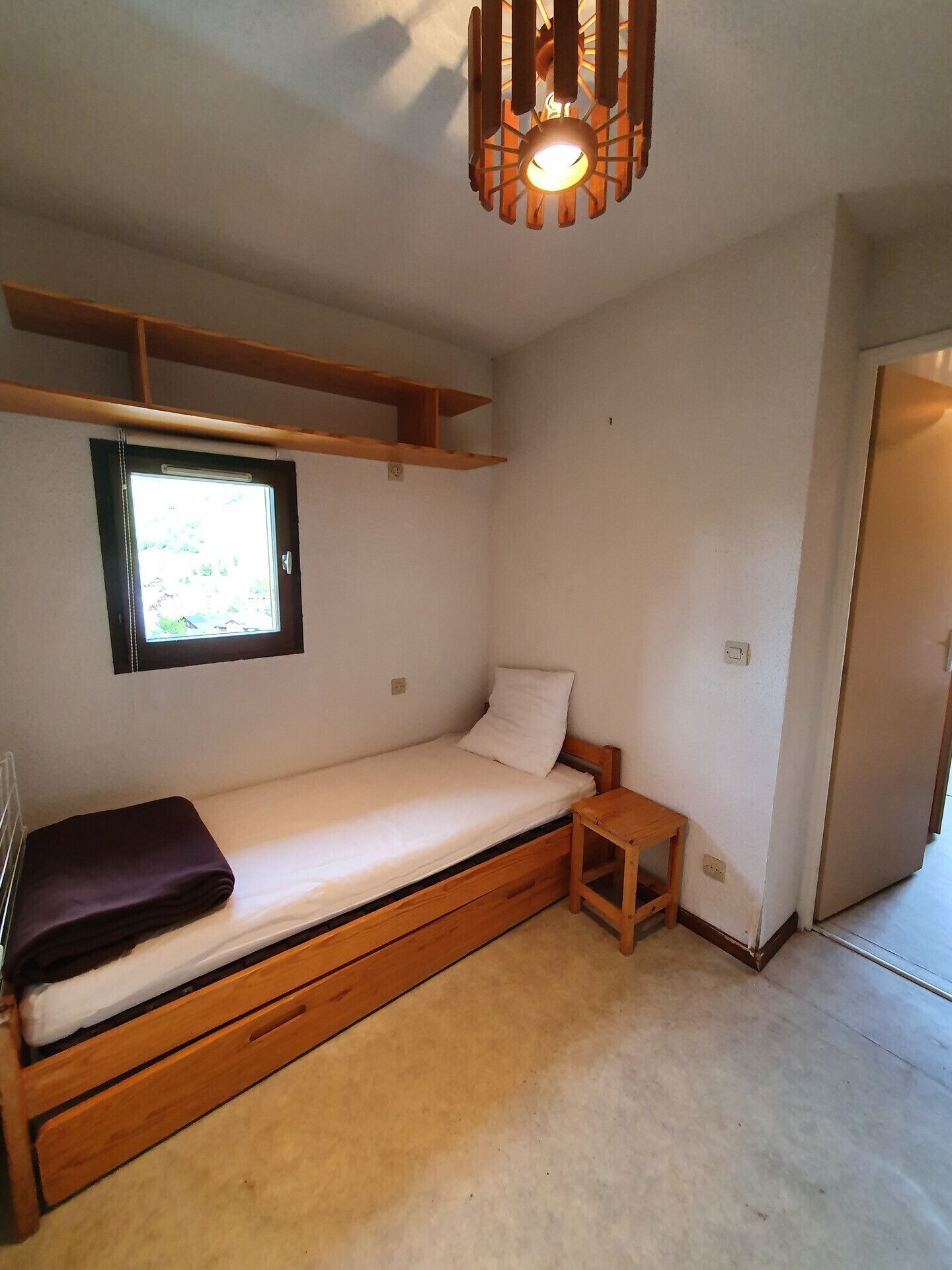 A small, single-bed room with a wooden bed frame, a small wooden nightstand, a window with a view, and a ceiling light fixture.