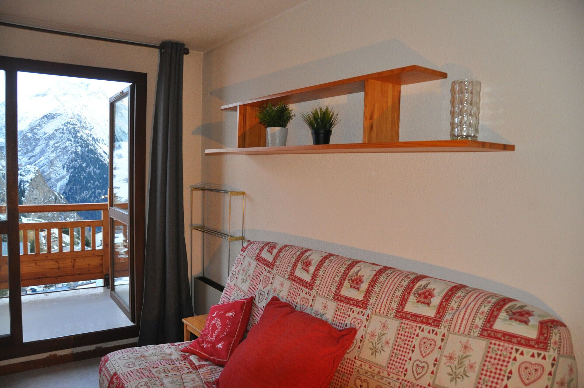 A cozy living room with a wooden sofa, a red cushion, and a patterned throw. A wooden shelf with decorative items is mounted on the wall.