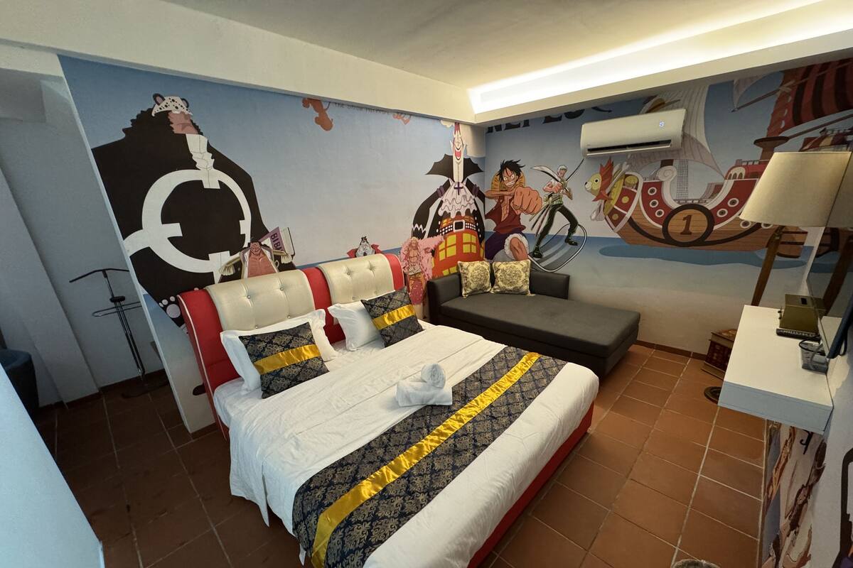 A hotel room with a large bed, a sofa, a wall mural, and a small table with a lamp.