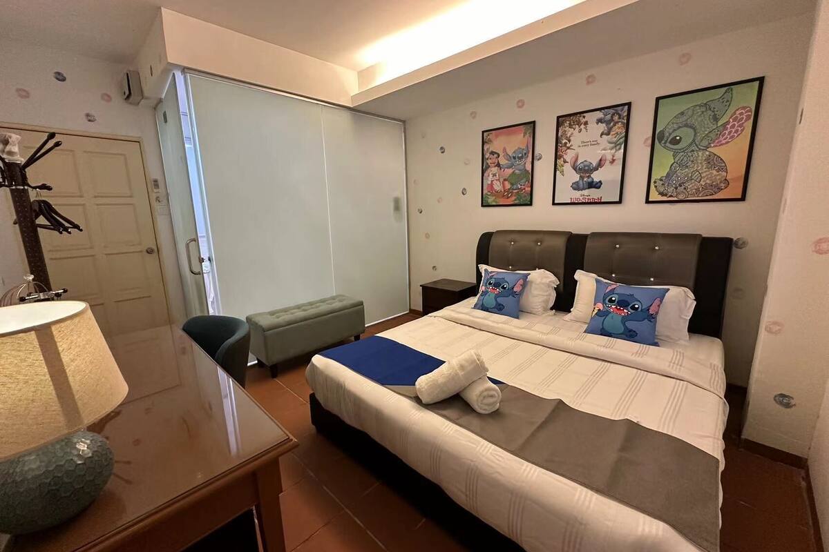 A hotel room with a large bed, a desk with a lamp, a bench, and three wall-mounted pictures.