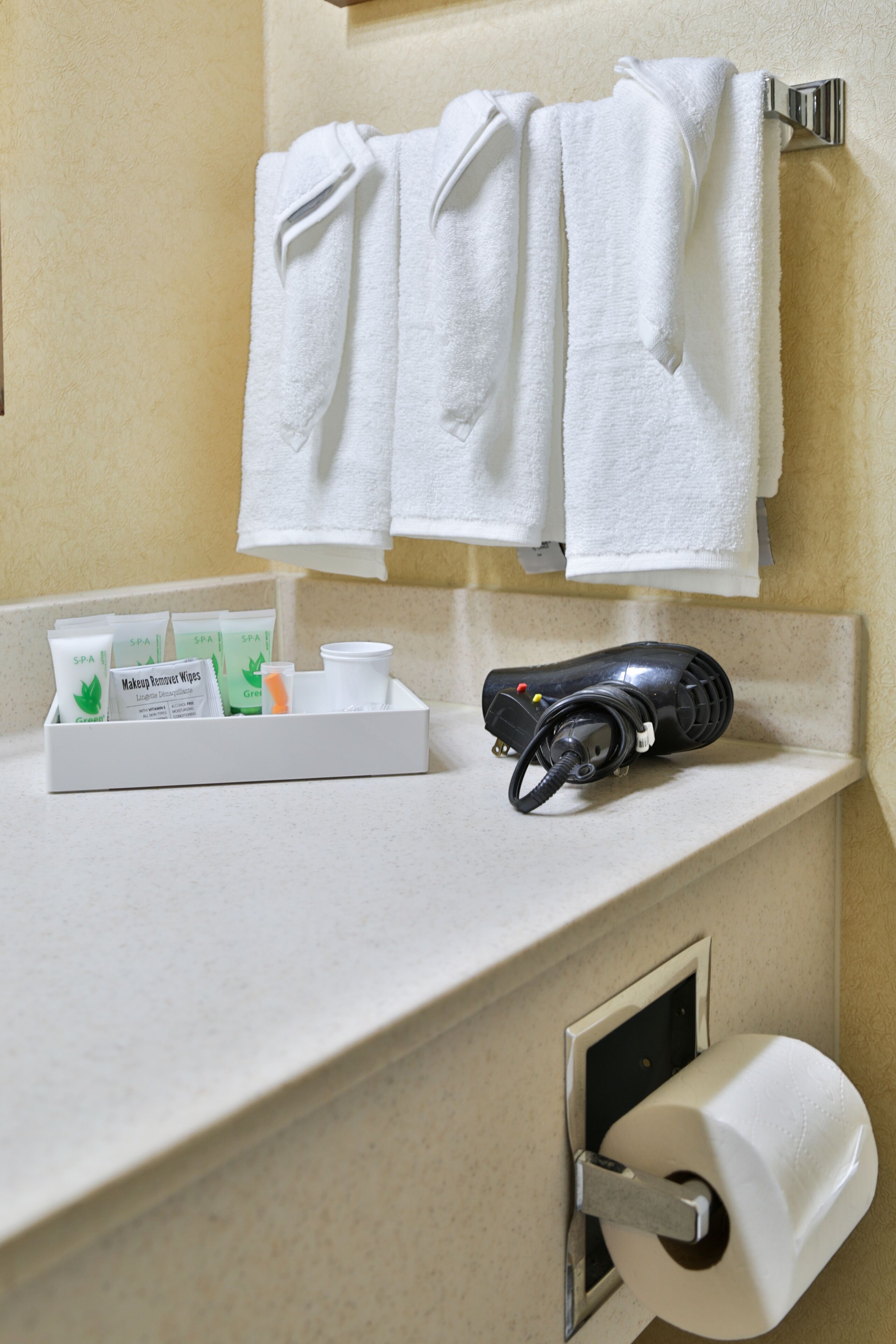 A bathroom with a towel rack holding three white towels, a hairdryer, and a tray with toiletries including makeup wipes.