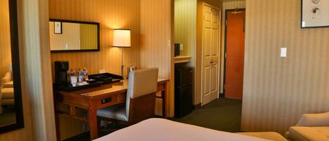 A hotel room with a bed, a desk with a chair, a lamp, a mirror, a refrigerator, and a door leading to another room.