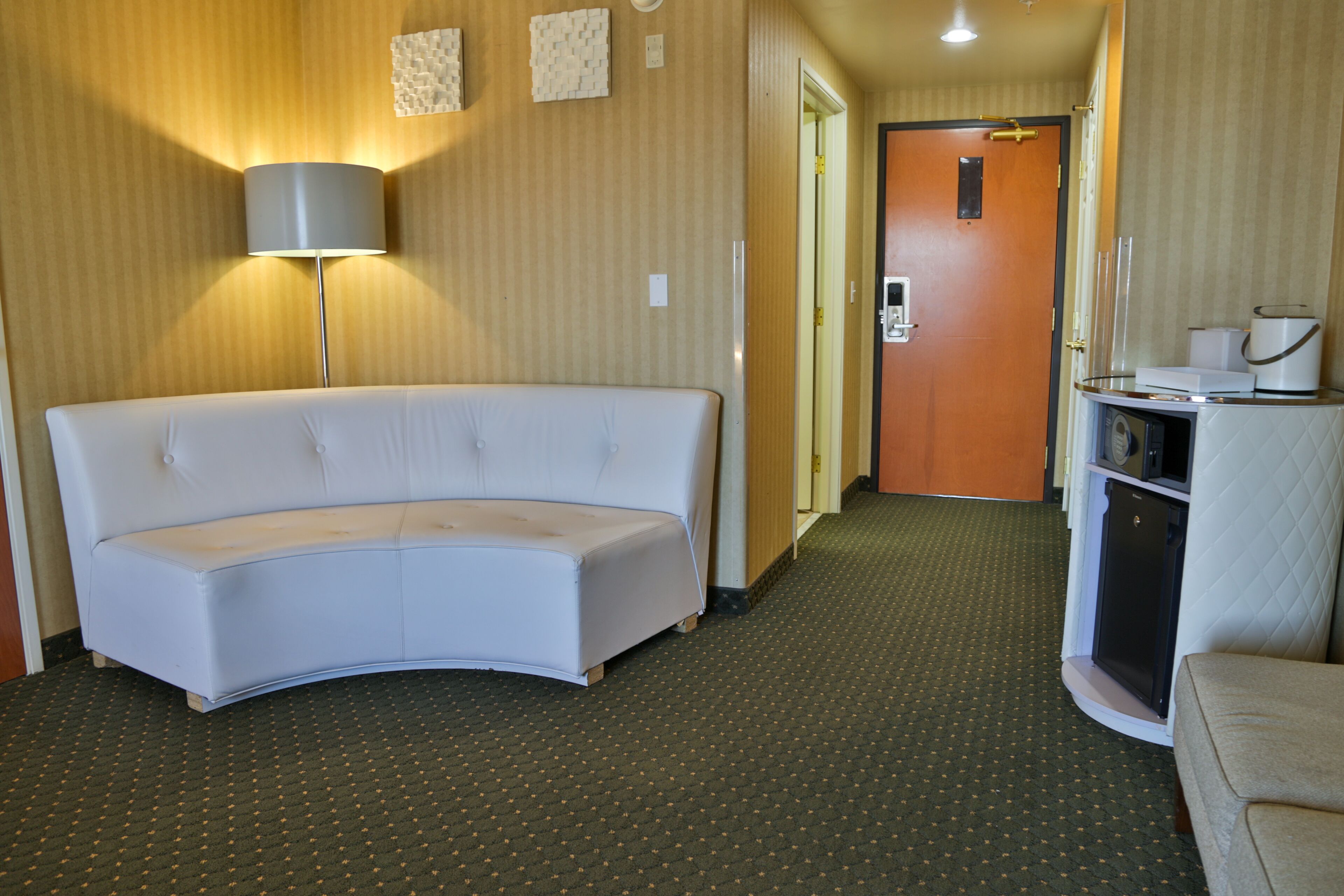 A hotel room with a white curved sofa, a small kitchenette, a lamp, and a door leading to another room.