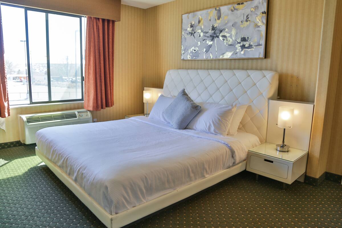 A hotel room with a large bed, a nightstand with a lamp, a window with curtains, and a wall-mounted abstract painting.