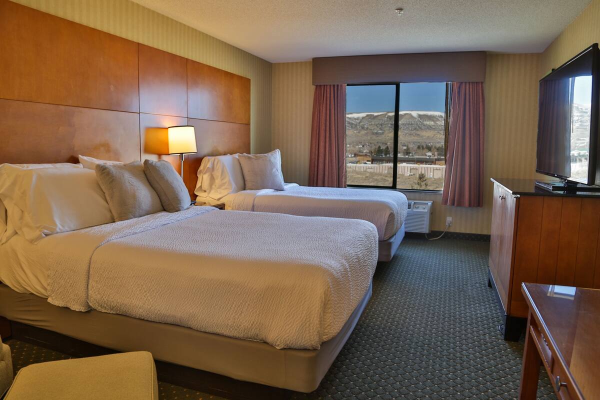 A hotel room with two beds, a desk, a lamp, and a large window with a mountain view.
