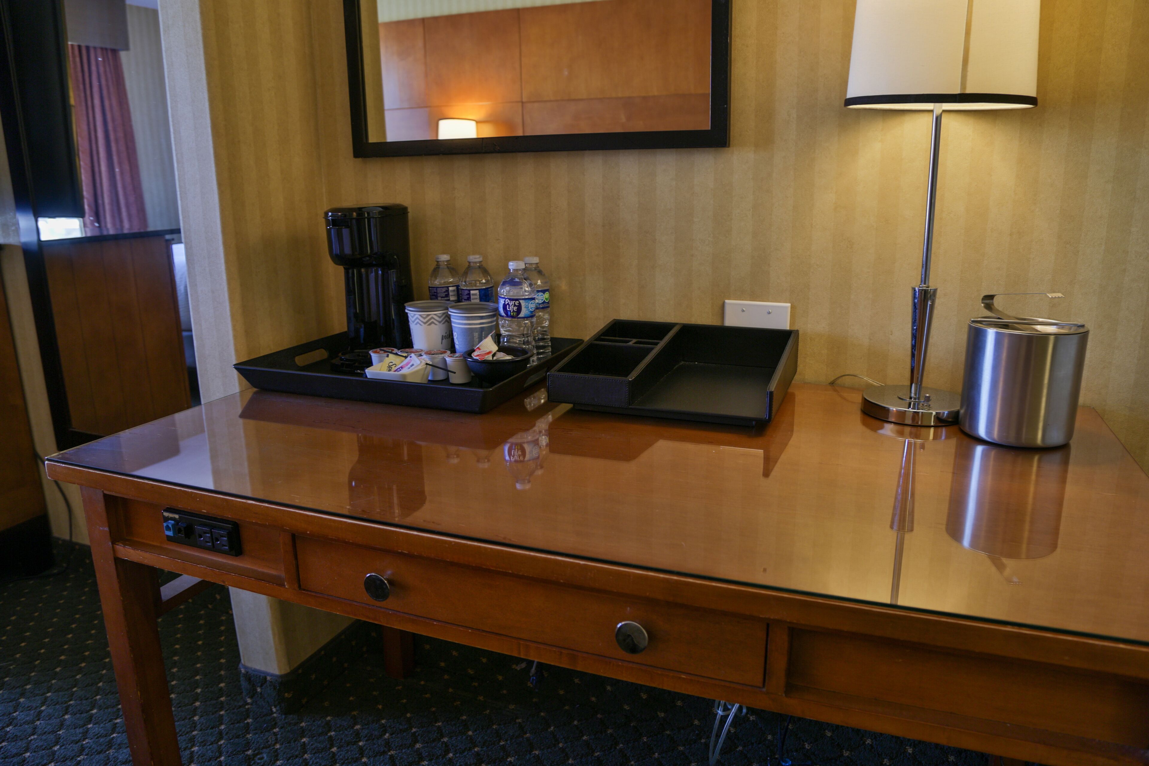 A hotel desk with a coffee maker, water bottles, a lamp, and a trash can.