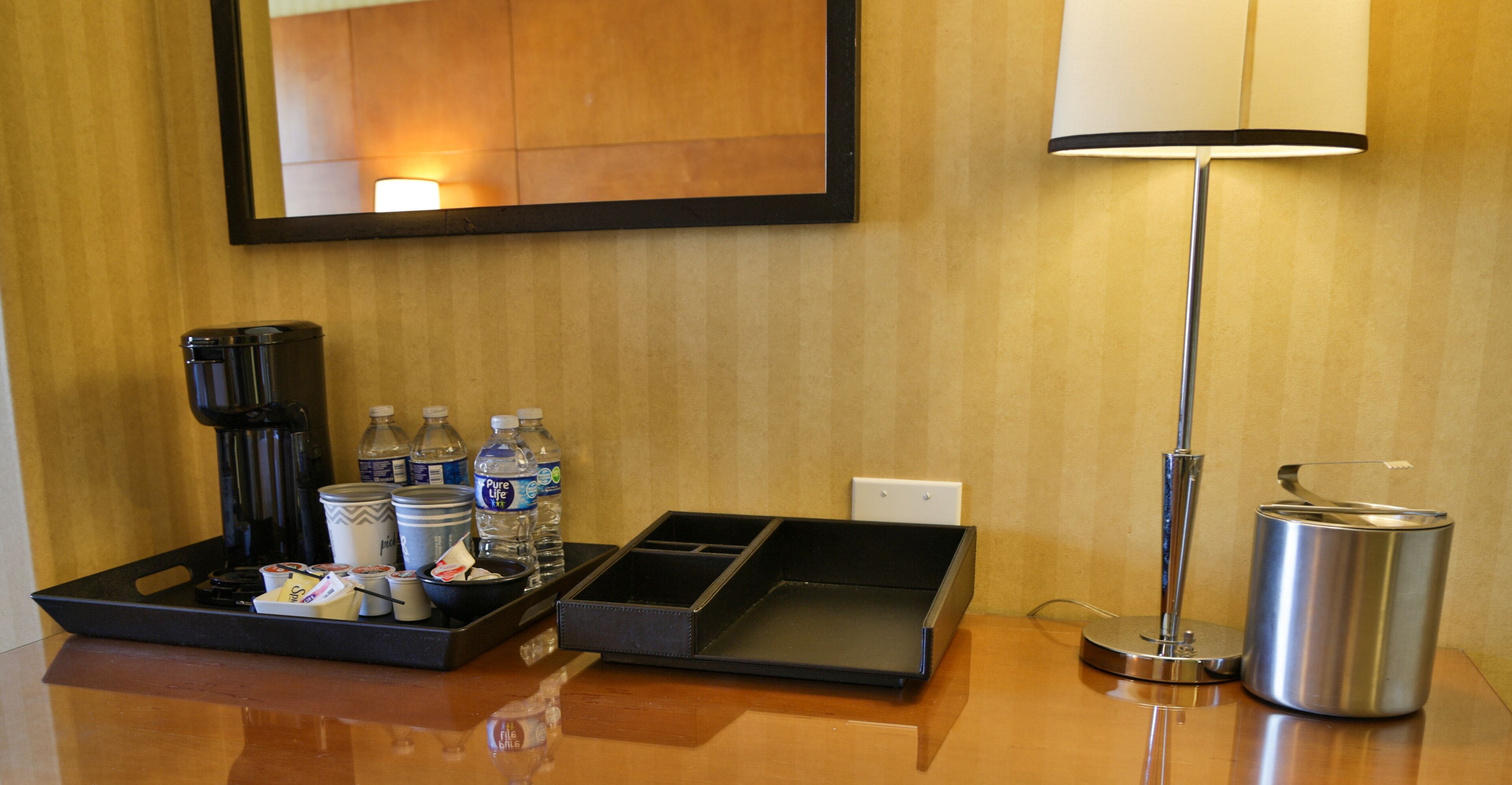 Un bureau dans une chambre d’hôtel, avec une machine à café, des bouteilles d’eau, un plateau avec des en-cas, une lampe et une poubelle.