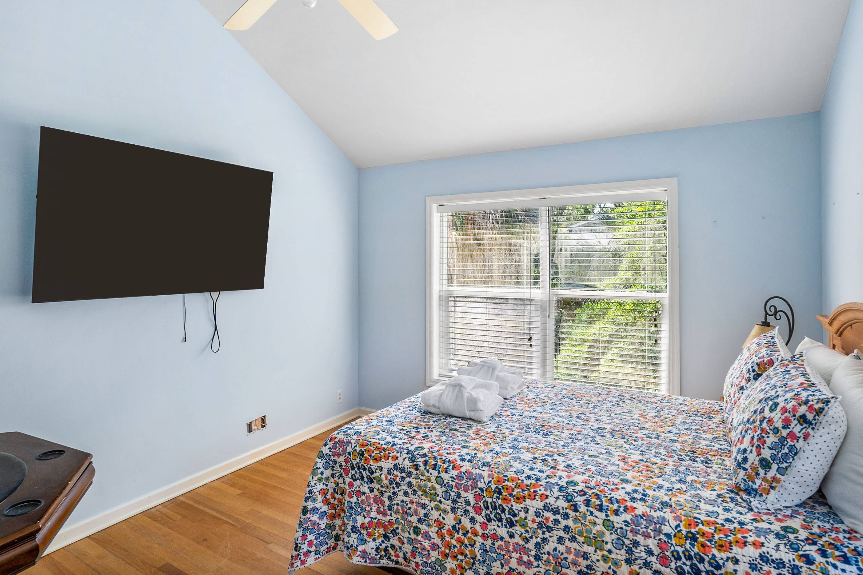 Un dormitorio con cama, televisor de pantalla plana montado en la pared, ventana con persianas y suelo de madera.