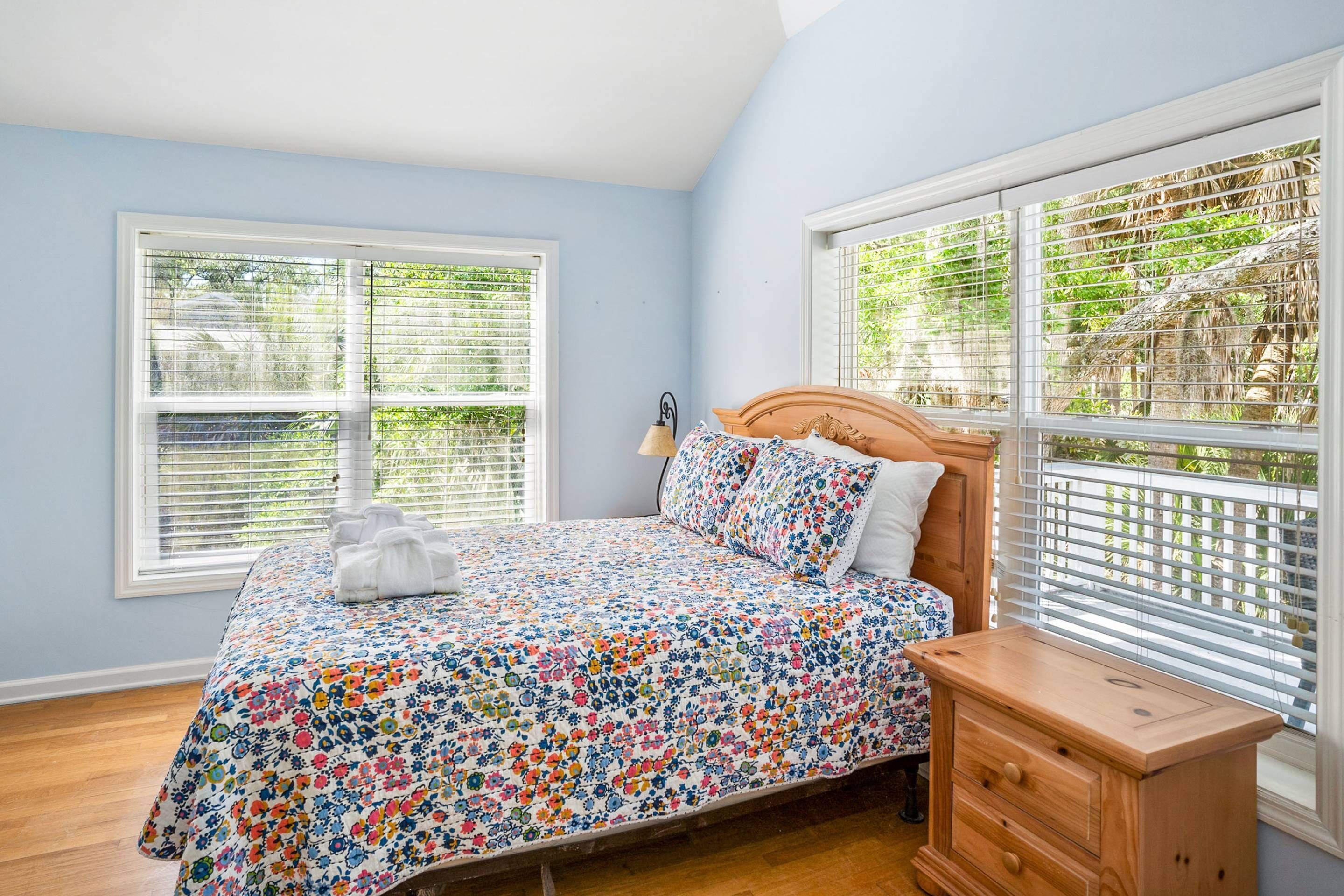 A bedroom with a floral bedspread, a wooden nightstand, and a window with blinds.