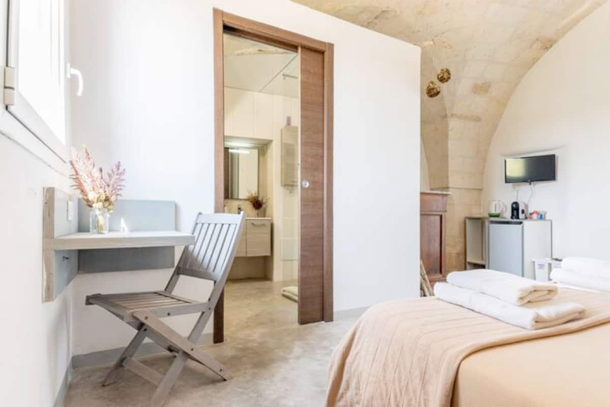 Une chambre à coucher comprenant un lit, un bureau, une chaise, un téléviseur et une salle de bain visible à travers une porte ouverte.