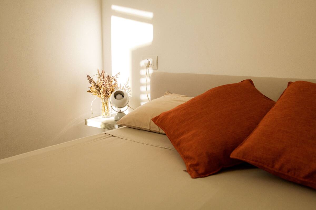 Un lit bien fait, avec deux oreillers, une table de chevet surmontée d’une lampe et un vase contenant des fleurs séchées.