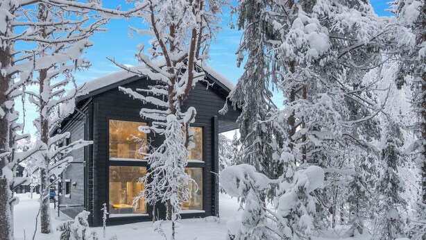 Una cabaña cubierta de nieve con un ventanal grande, rodeada de árboles cargados de nieve.