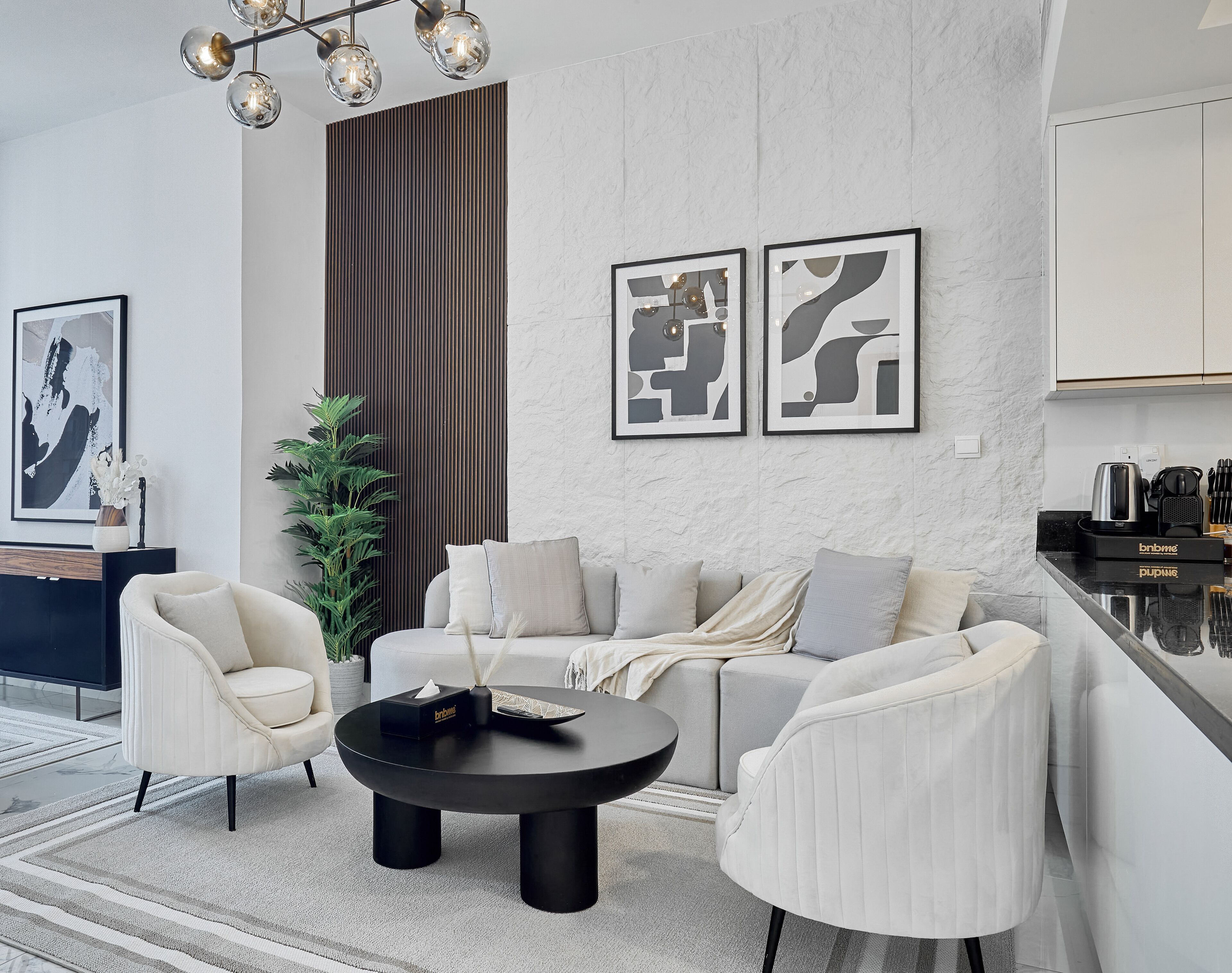 Un salon moderne avec une table basse ronde noire, deux fauteuils blancs, un canapé gris et une grande plante.