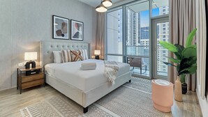 A modern hotel room with a large bed, bedside tables, a nightstand, a large window with a view of buildings, and a potted plant.