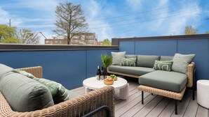 Eine Dachterrasse mit Korbmöbeln, einem grünen Sofa und einem runden Tisch mit Flaschen und einer Obstschale.