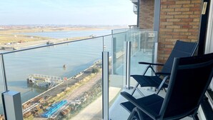 Ein Balkon mit Glasgeländer, der auf einen Fluss und eine Brücke blickt. Zwei schwarze Stühle und ein Tisch sind ebenfalls vorhanden.