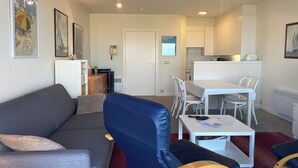 Ein modernes Wohnzimmer mit einem grauen Sofa, einem blauen Sessel und einem kleinen weißen Tisch. Es gibt einen weißen Essbereich mit Tisch und Stühlen. Die Wände sind weiß und es gibt eine Tür, die in eine Küche führt.