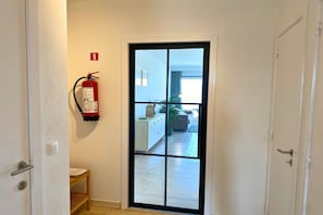 A door leading to a room with a fire extinguisher mounted on the wall, a visible fire exit sign, and a view of a living area with a sofa and a plant.