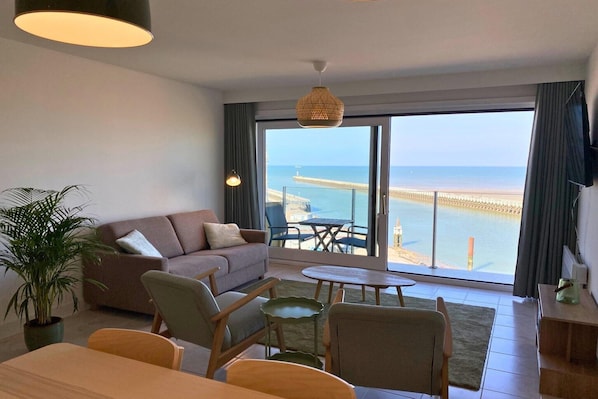 A modern living room with a sofa, chairs, a dining table, and a view of the beach through large windows.
