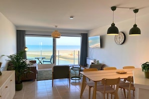 A modern living room with a dining area, large windows, a clock, and a view of the sea.