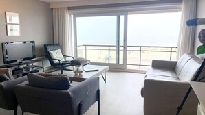 A modern living room with a television, seating, and a large sliding glass door leading to a balcony with a view of the beach. - Apartment in Nieuwpoort by Scenic Beaches (Nieuwpoort)