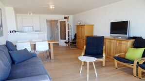 A modern living room with a grey sofa, a small round table, and a TV. There is a kitchen area with white cabinets and a dining table. - Apartment in Nieuwpoort by Scenic Beaches (Nieuwpoort)