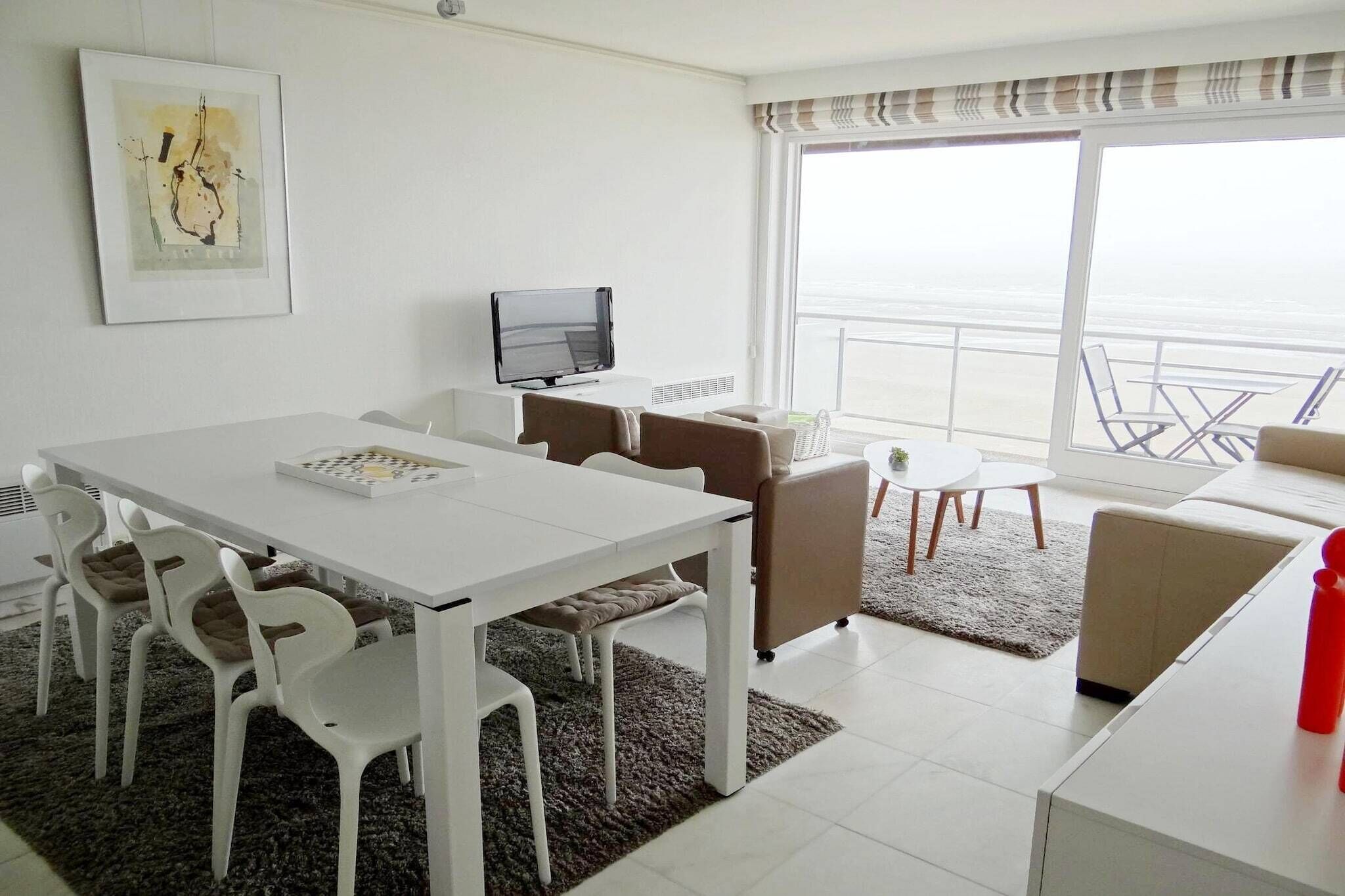 A modern hotel room with a white dining table, chairs, a flat-screen TV, a sofa, and a view of the ocean.
