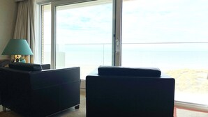A room with a large sliding glass door leading to a balcony with a view of the sea. There are two black sofas and a teal floor lamp. - Apartment in Nieuwpoort near the Beach (Nieuwpoort)