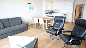 Uma sala de estar moderna com um sofá cinzento, uma mesa de jantar com cadeiras e uma poltrona de couro preto.