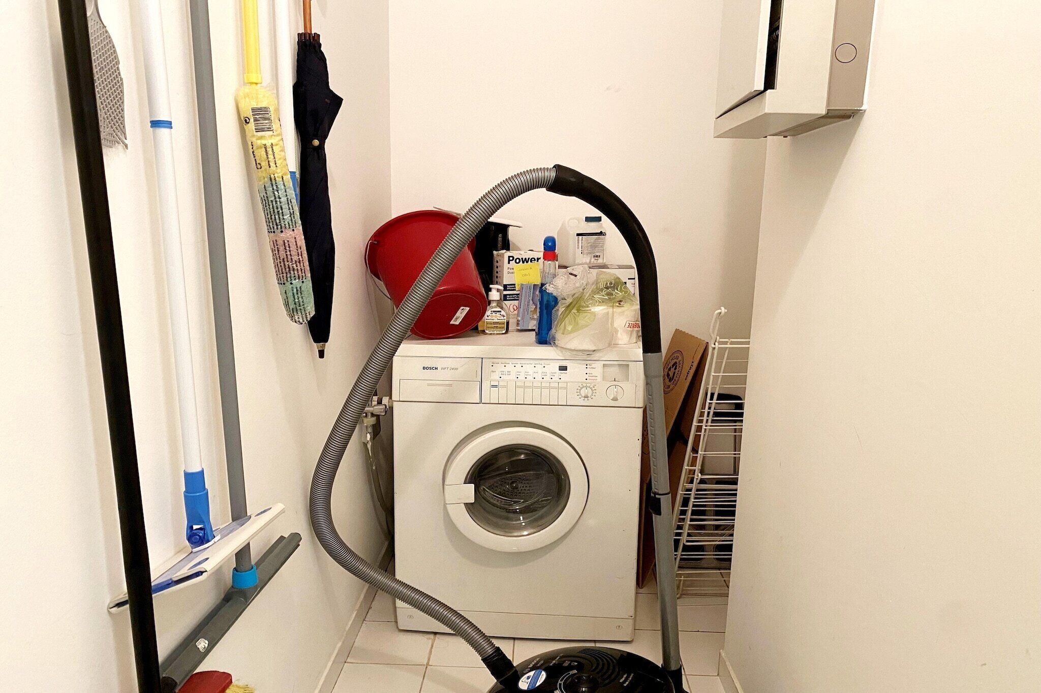 Une petite buanderie équipée d’un lave-linge à chargement frontal, d’un aspirateur et de divers produits de nettoyage.