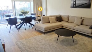 A modern living room with a beige sofa, a round dining table with chairs, a coffee table, and a large window offering a view of a cityscape. - Apartment in Nieuwpoort near the Beach (Nieuwpoort)