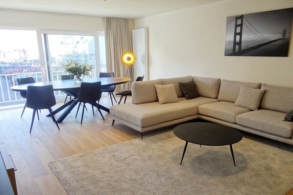 Ein modernes Wohnzimmer mit einem beigen Sofa, einem runden Esstisch mit Stühlen, einem Couchtisch und einem großen Fenster mit Blick auf eine Stadtlandschaft.