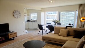 A modern living room with a sofa, a coffee table, and a TV. There is a dining area with chairs and a table, and a kitchen in the background. - Apartment in Nieuwpoort near the Beach (Nieuwpoort)