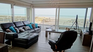 Un moderno salón con un sofá de cuero negro, una silla de oficina negra y una mesa de comedor. Amplias ventanas ofrecen vistas a la playa y al océano.