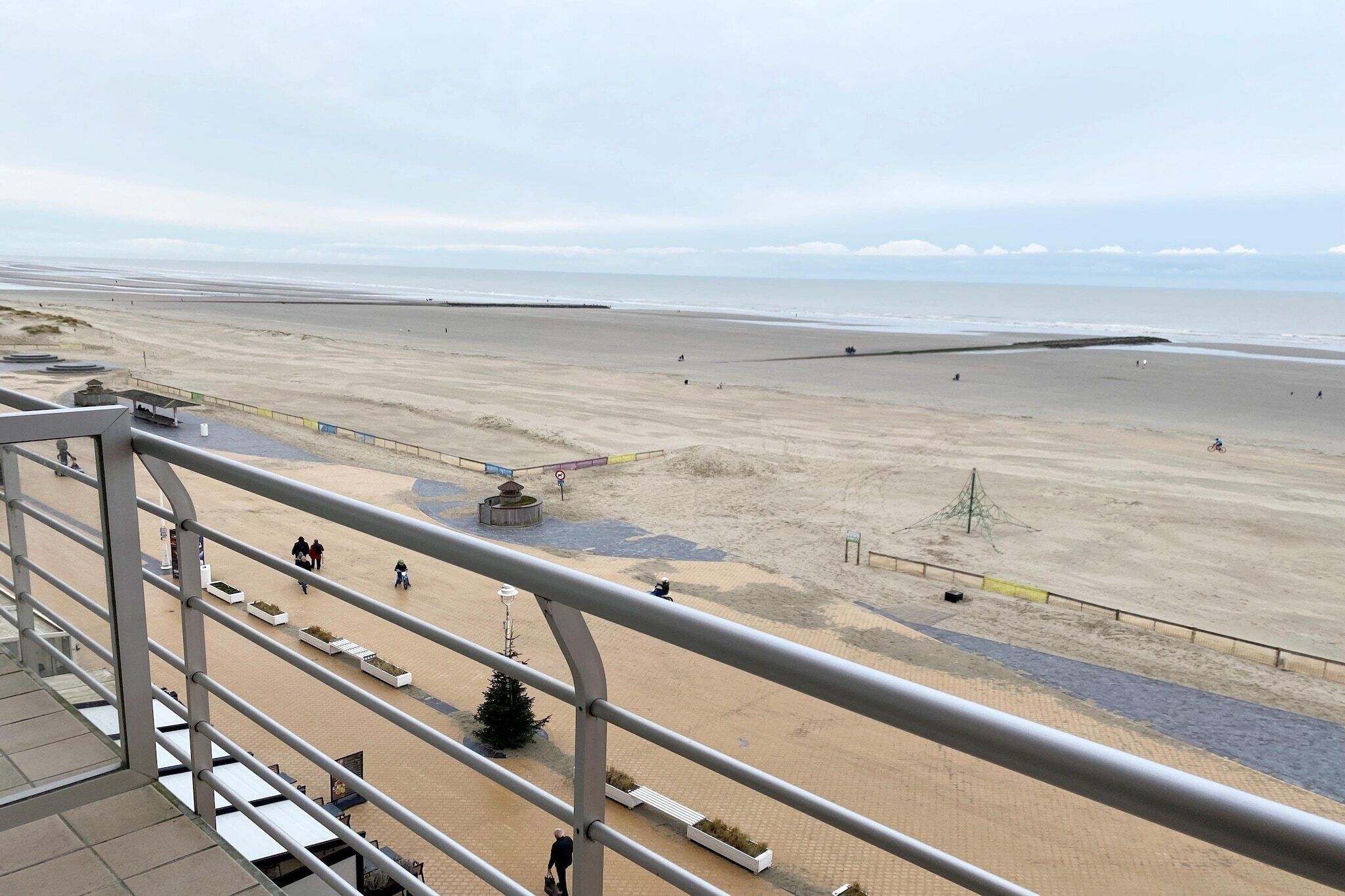 Vista de una playa desde un balcón con barandilla, playa de arena y algunas personas caminando.