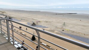 Vista de una playa desde un balcón con barandilla, playa de arena y algunas personas caminando.