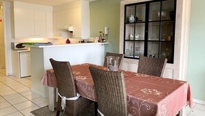 Un coin repas avec une table dressée pour quatre personnes, entourée de chaises en osier, une cuisine en arrière-plan et un buffet dont les portes en verre exposent de la vaisselle.