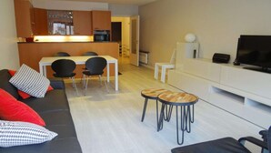 Ein modernes Wohnzimmer mit einer Couch, einem Esstisch mit Stühlen, einem Fernseher und einer Küche im Hintergrund.
