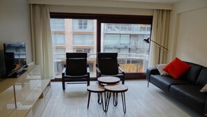 Ein modernes Wohnzimmer mit einem schwarzen Sofa, einem Couchtisch, zwei Sesseln, einem Fernseher und einem großen Fenster mit Blick auf ein Gebäude.