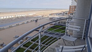 A balcony with a view of a beach, a parking area, and a building with a curved facade. - Apartment in Nieuwpoort with Ocean Views (Nieuwpoort)
