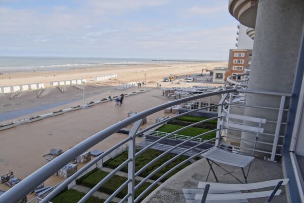 A balcony with a view of a beach, a parking area, and a building with a curved facade.