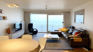 A modern living room with a TV, sofa, chairs, and a balcony with a view. - Apartment in Nieuwpoort with Sea Views (Nieuwpoort)