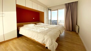 Une chambre moderne avec un grand lit, un plancher en bois, un mur d’accent rouge et une vue dégagée sur l’extérieur grâce à une grande fenêtre.