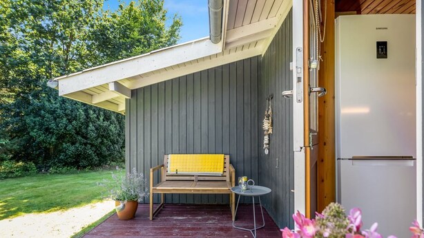 Une terrasse en bois avec un banc et une petite table, un réfrigérateur et une porte donnant sur une pièce.