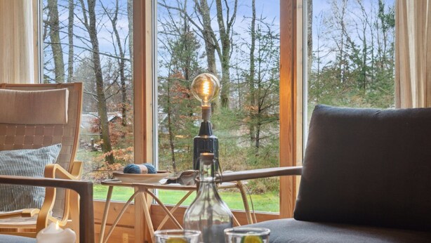 A cozy seating area with a view of trees through large windows, a candle, and a table with glasses and a remote control. - "Sofja" - 175m to the fjord by Interhome (Hadsund)