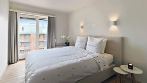 A hotel room with a large bed, two bedside tables, a nightstand, and a view of buildings through the window. - Apartment Nieuwpoort near Beach and Marina (Nieuwpoort)