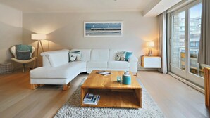 A modern living room with a white sofa, a wooden coffee table, and a large window with a view of buildings. - Apartment Nieuwpoort near Beach and Marina (Nieuwpoort)