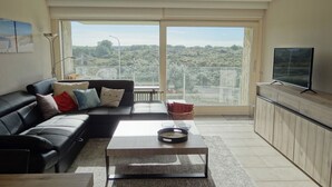 Un salon moderne avec un canapé en cuir noir, une table basse et un téléviseur posé sur un meuble en bois. Une grande fenêtre offre une vue sur un paysage verdoyant.